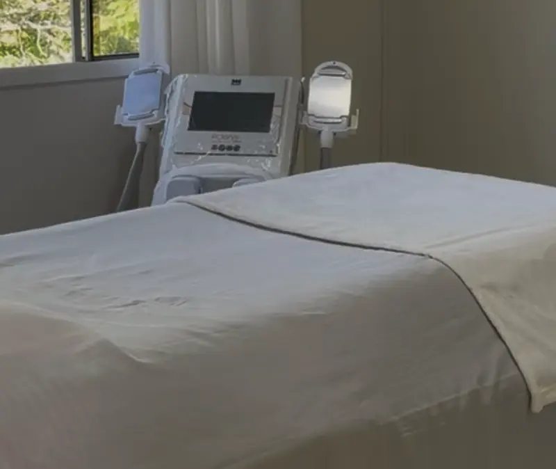 Sala de tratamento corporal com maca e aparelhos, iluminada pela janela